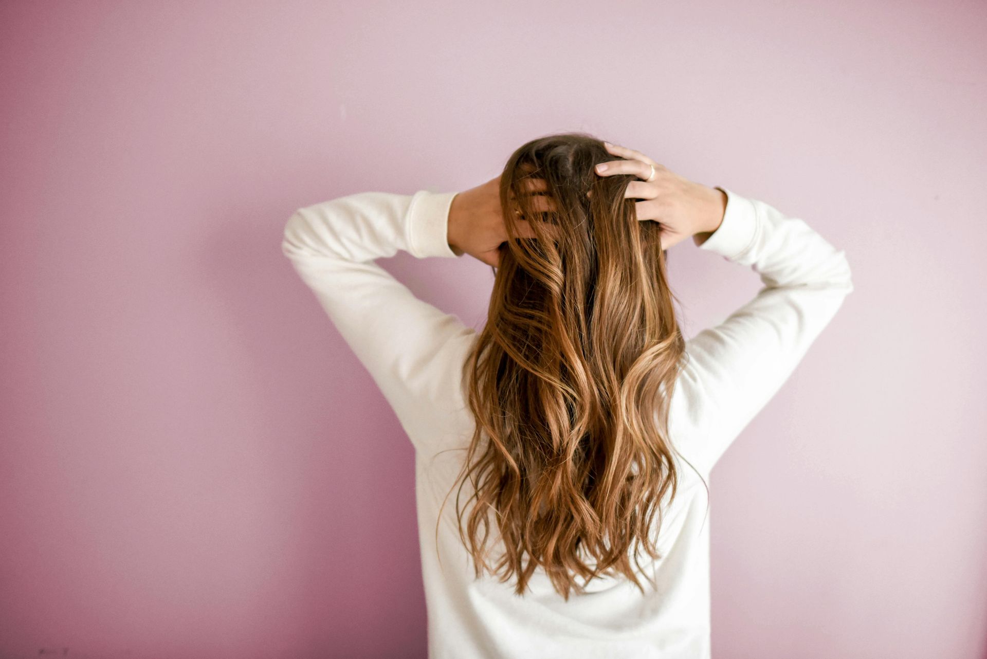 Woman with long, wavy brown hair, hands in hair, wearing white sweater, standing against a pink wall.