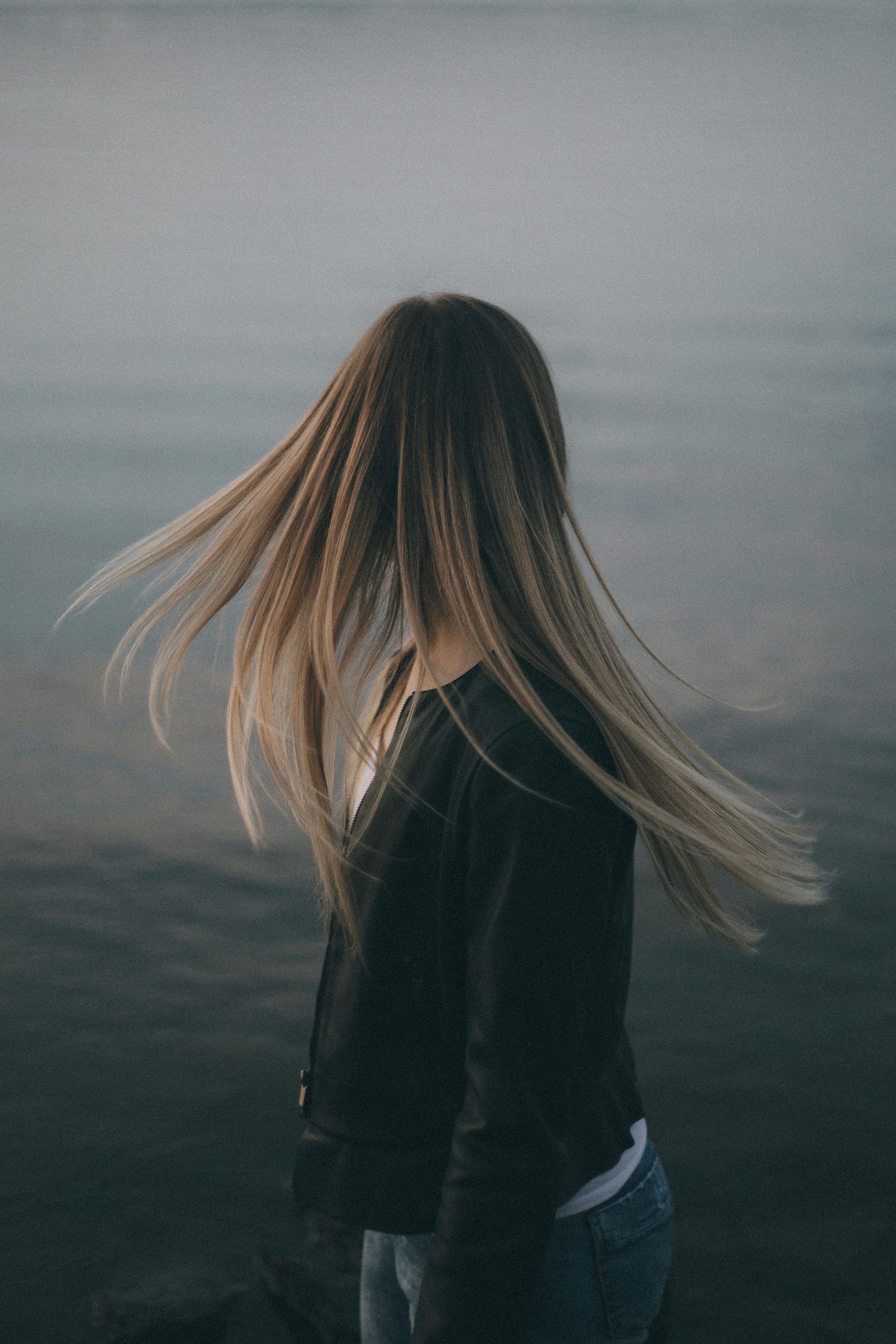 Woman with long hair twirling near water, wearing a black jacket and jeans.