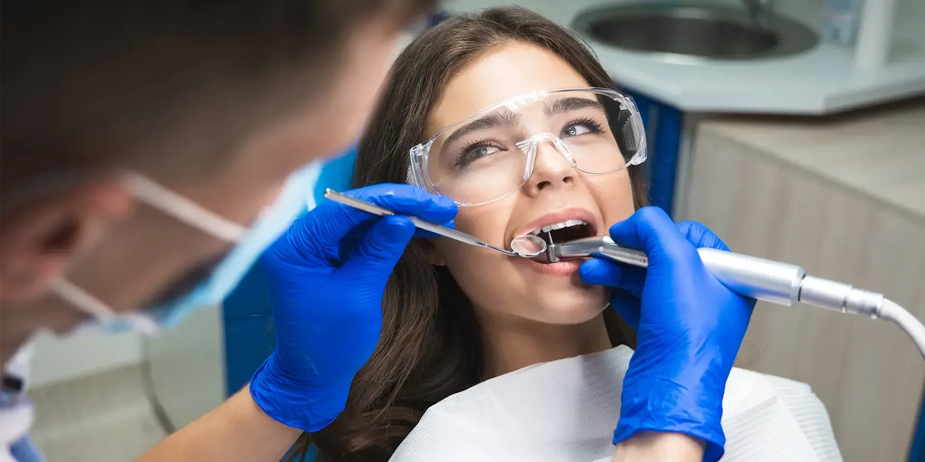 A dentist wearing blue gloves and a face mask uses tools to examine a patient’s teeth in a clinical setting.