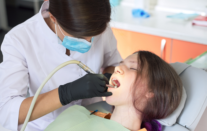 A dentist wearing a mask and black gloves works on a patient's teeth in a clinical setting.