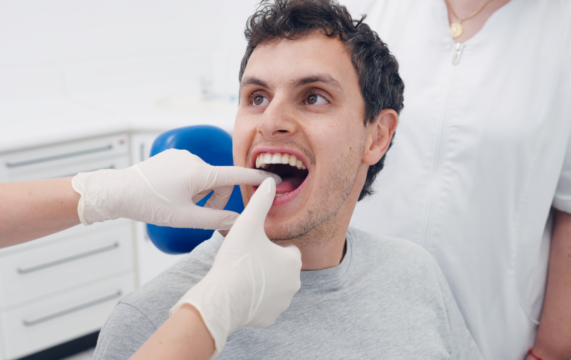 A dentist wearing gloves performs an examination on a patient's teeth in a clinical office setting.