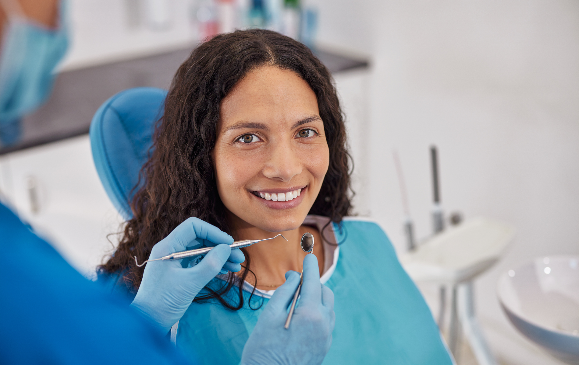 A dental hygienist wearing blue gloves examines the teeth of a smiling patient in a teal bib at a dental clinic.
