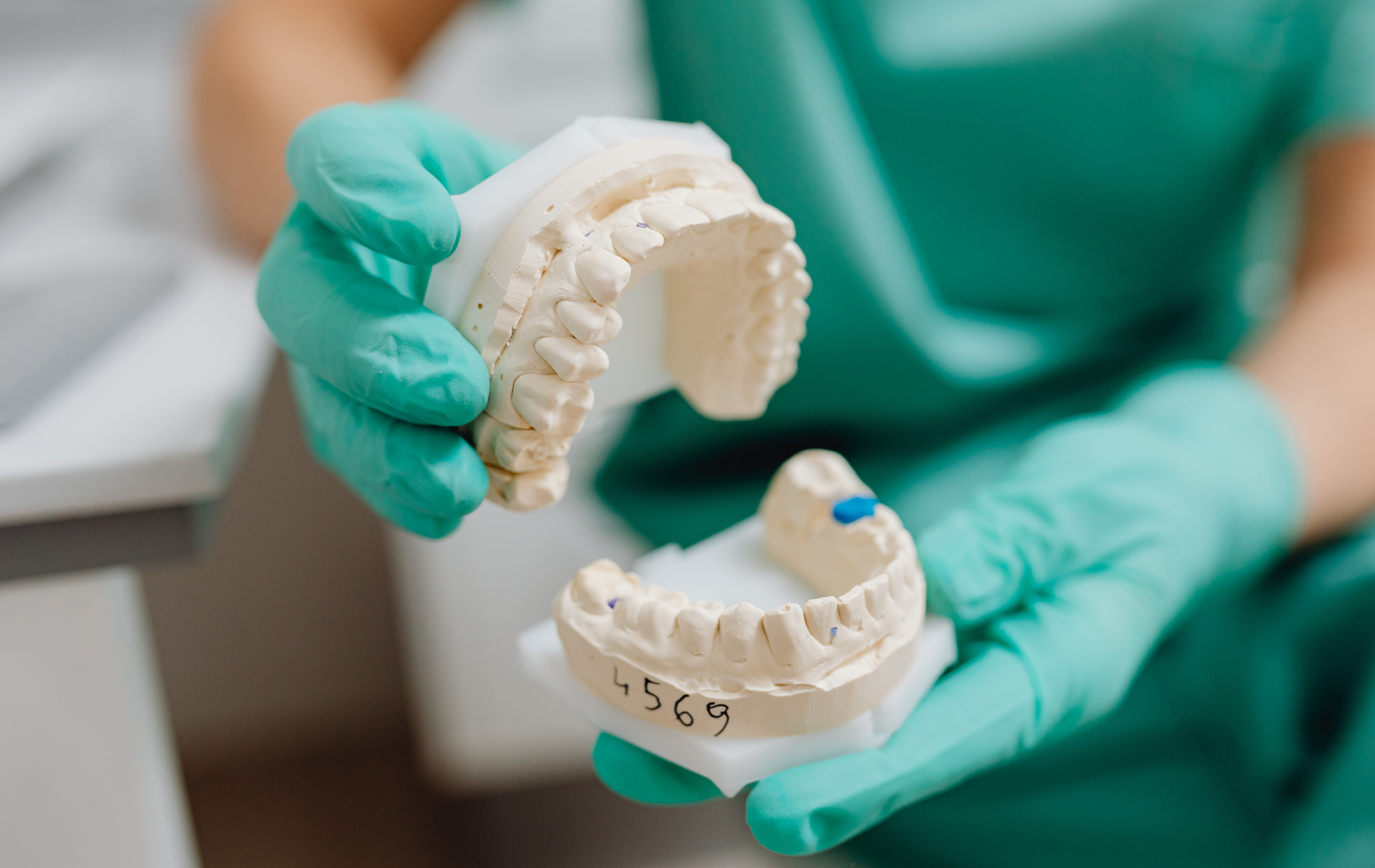 A person in teal gloves holding plaster dental models of upper and lower teeth in a clinical setting.