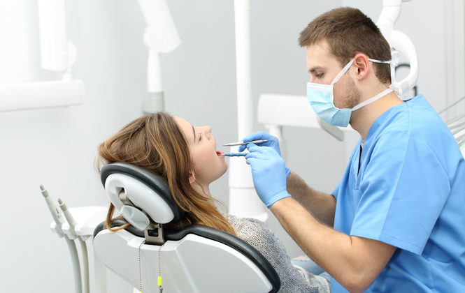 A masked dental professional in blue scrubs performs an examination on a patient reclined in a dental chair.