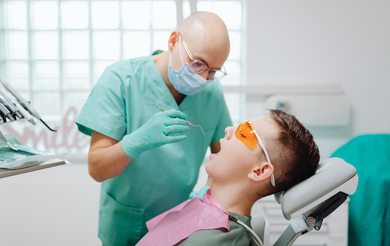 A dentist in scrubs and a mask examines a patient wearing protective orange glasses in a dental chair.