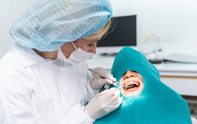A dentist wearing personal protective equipment performs a checkup on a patient wearing braces and a green dental bib.
