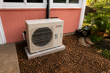 Outdoor air conditioner unit beside a pink house wall and window, on gravel near plants