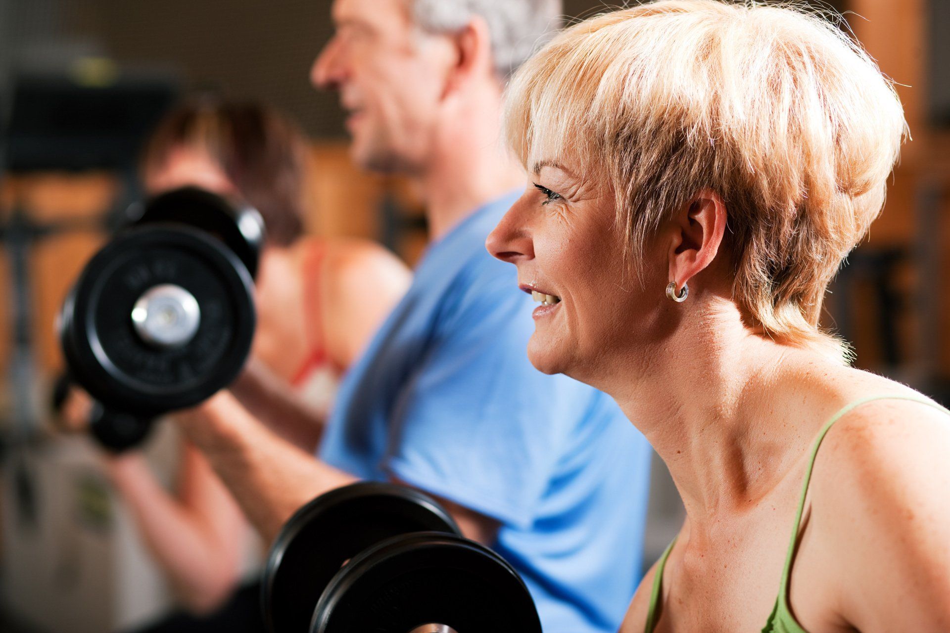People lifting weights in a gym; smiling woman in green tank top, man in blue shirt, weights, setting blurred.