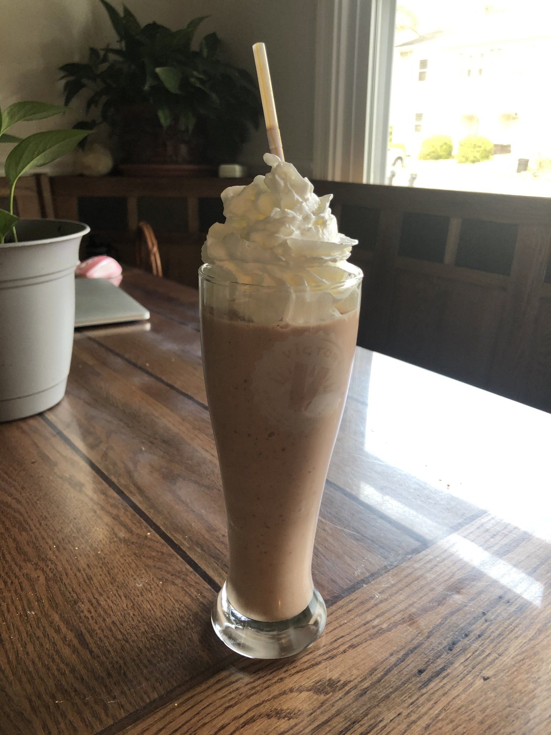 Chocolate milkshake in a tall glass, topped with whipped cream and a straw, on a wooden table.