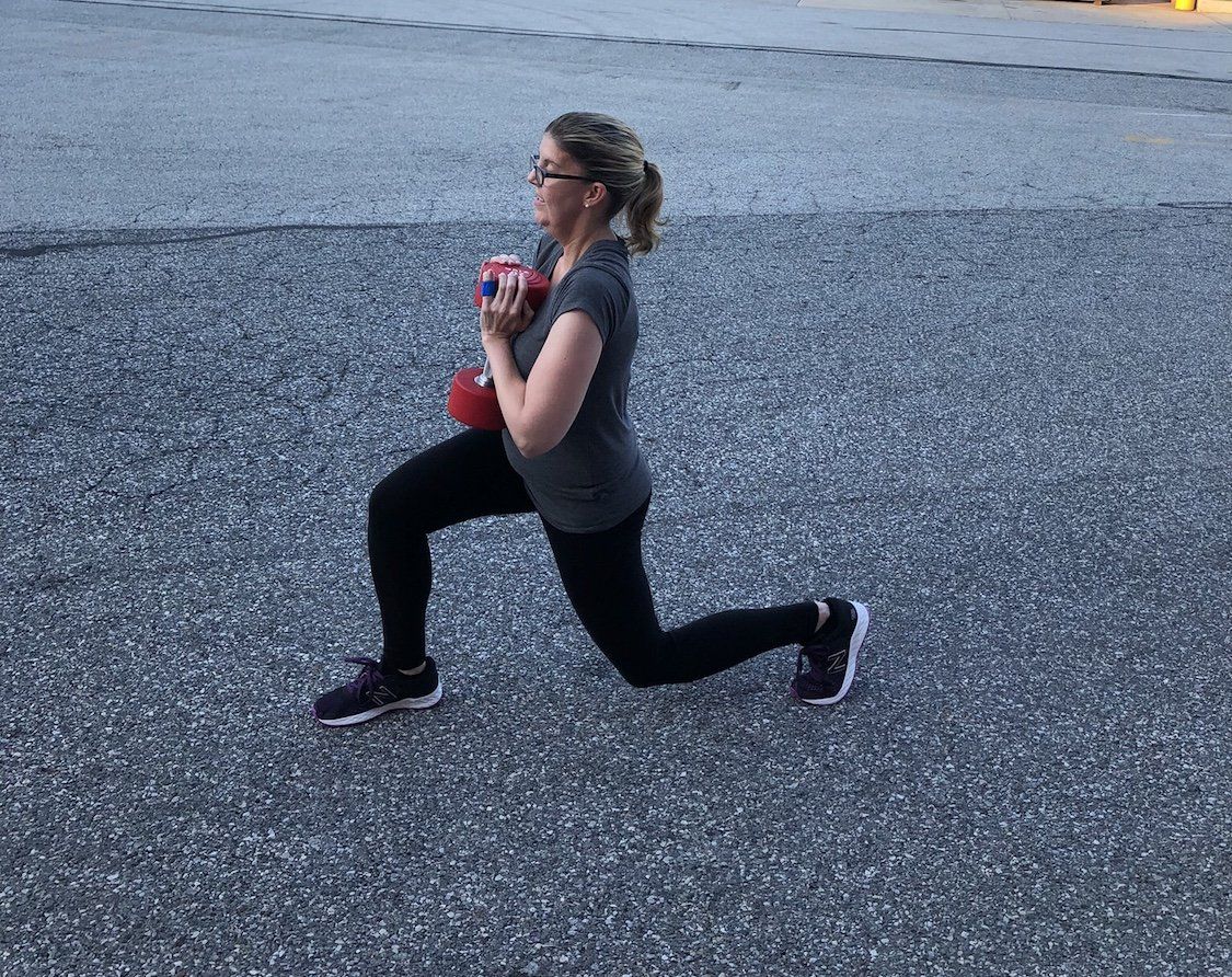 Woman in activewear performs a lunge while holding a weight outside.
