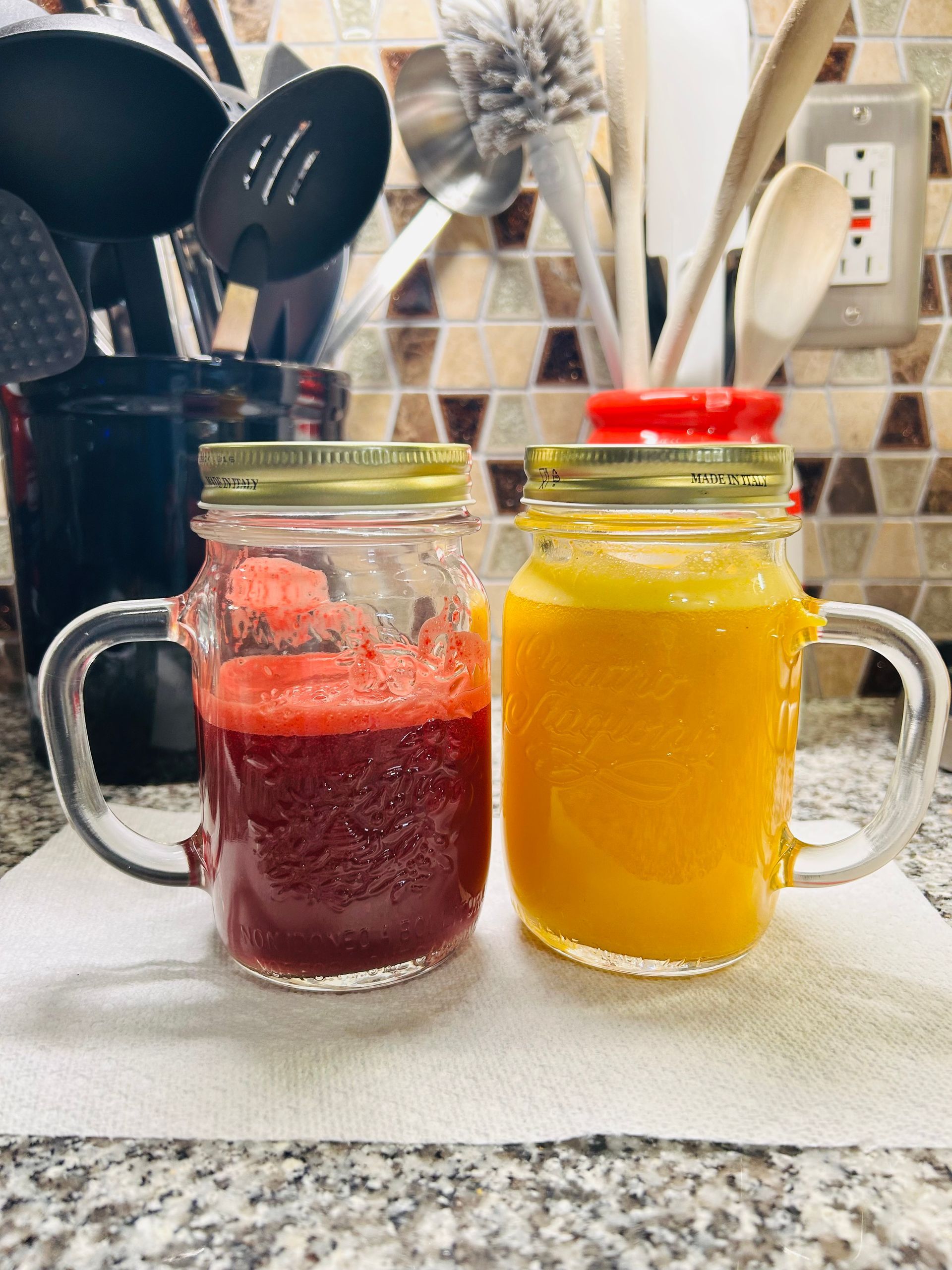 Two mason jar mugs with colorful juices; red and yellow, on a countertop, with kitchen utensils in the background.