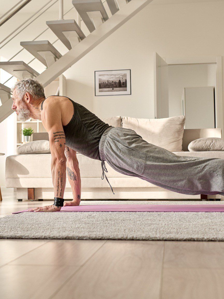 Man in a plank position on a yoga mat indoors; stairs and couch in the background.