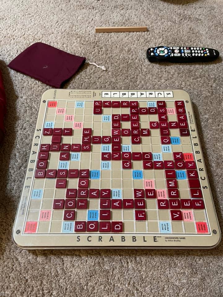 Scrabble board with played tiles on a beige surface, burgundy bag, wood rack, and remote control on a carpet.