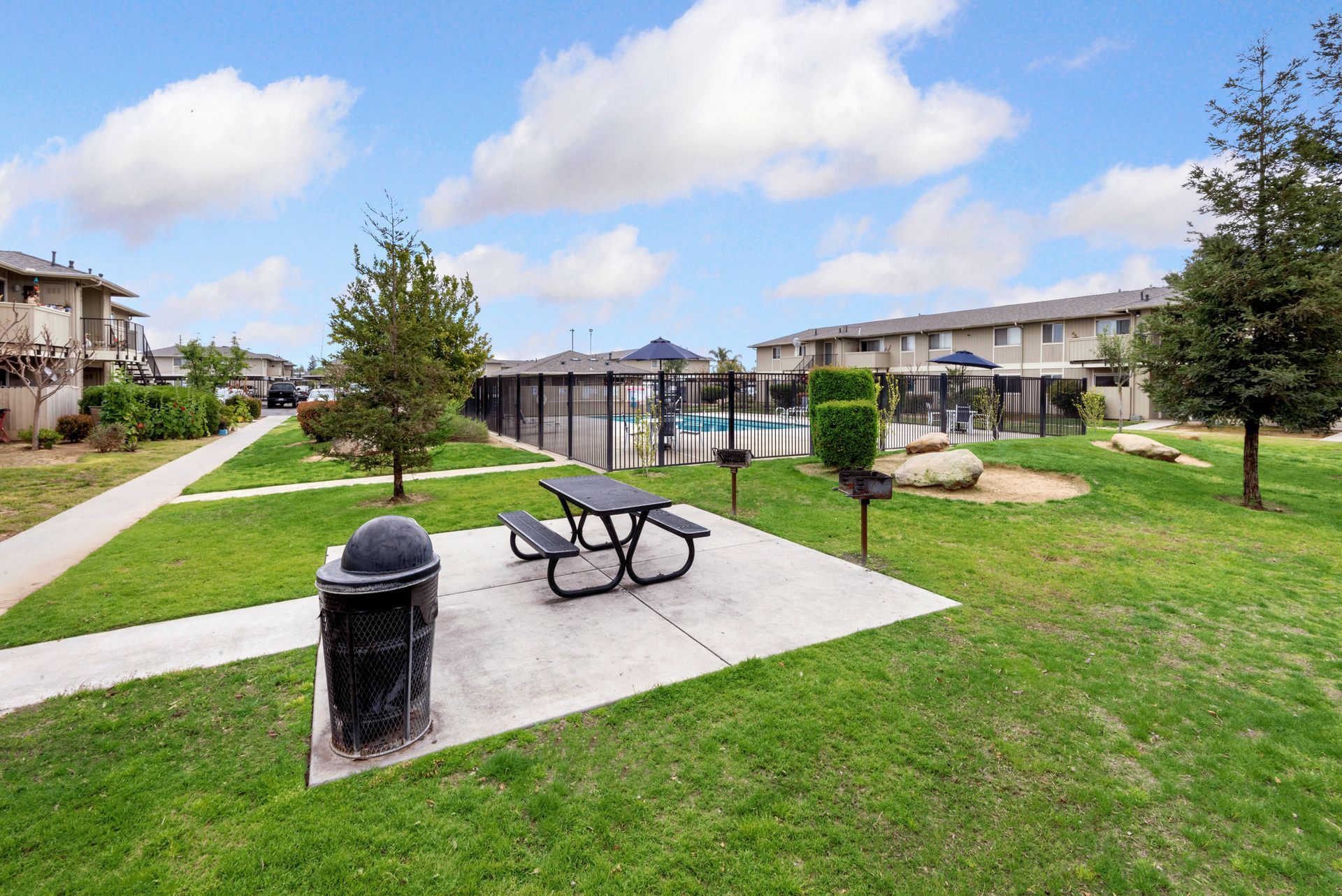 Park area with picnic table, trash can, pool, and apartment buildings on a sunny day.