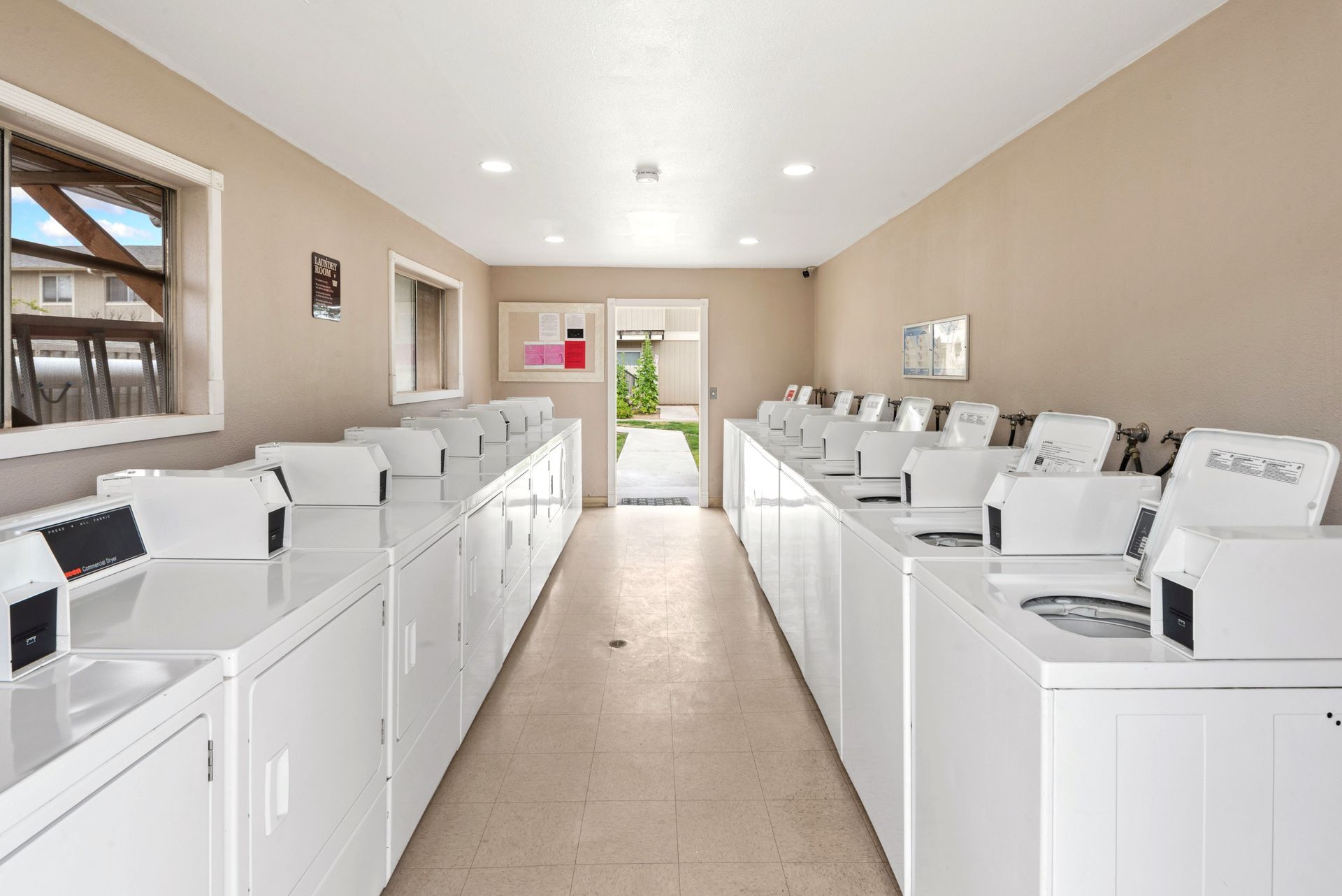 A laundry room with rows of white washing machines and dryers, a window, and a door leading outside.