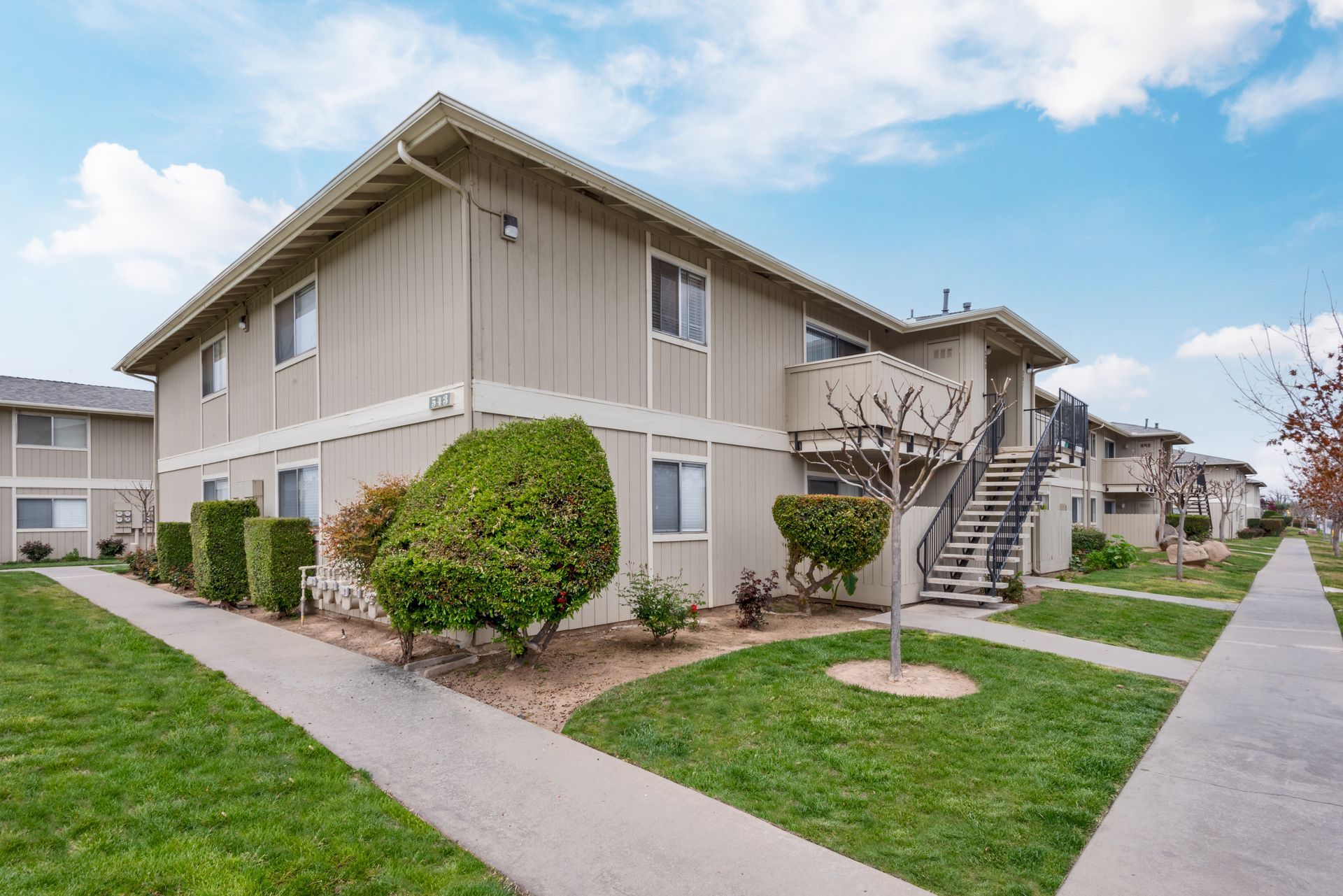 Two-story apartment building with beige siding, stairs, green lawn, and blue sky.