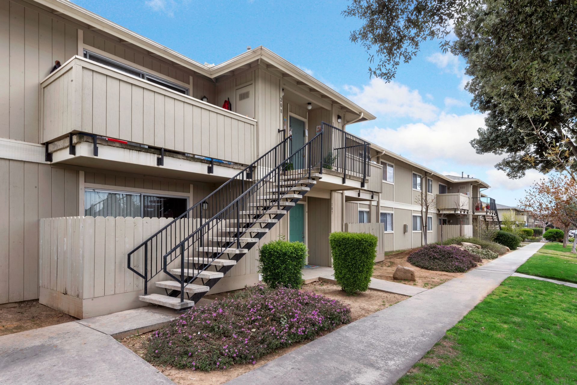 Apartment building exterior with stairs leading to upper units, beige siding, and landscaping along a sidewalk.