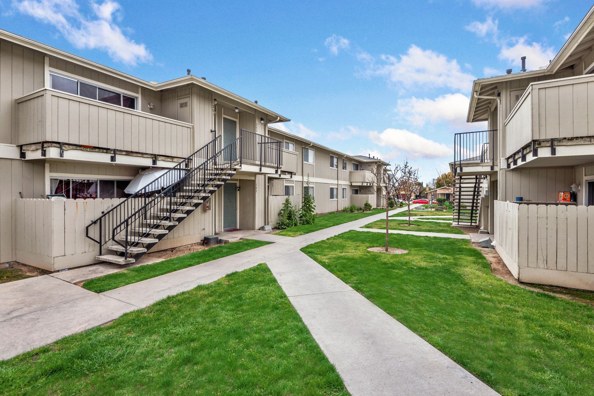 Apartment complex exterior with walkways, stairs, and green grass under a blue sky.