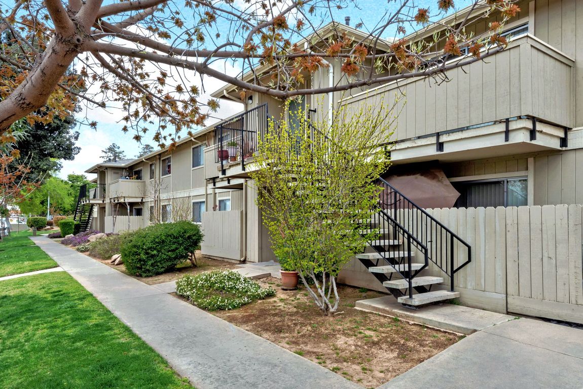Apartment complex with beige siding, stairs, and a walkway bordered by green grass and bushes.