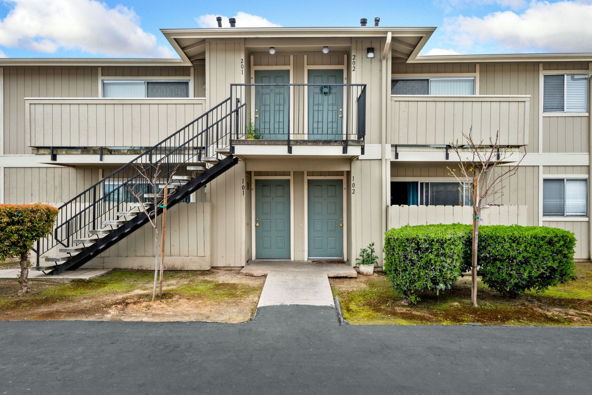 Two-story apartment building with teal doors and black exterior staircase. Beige siding, cloudy sky.