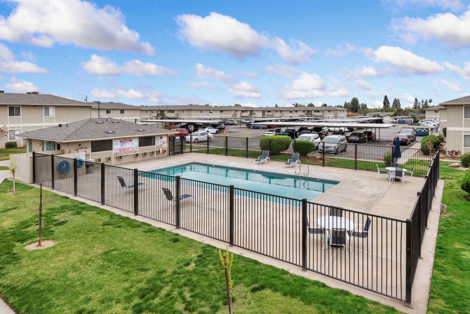 Apartment complex pool surrounded by a black fence, with cars under carports in the background.
