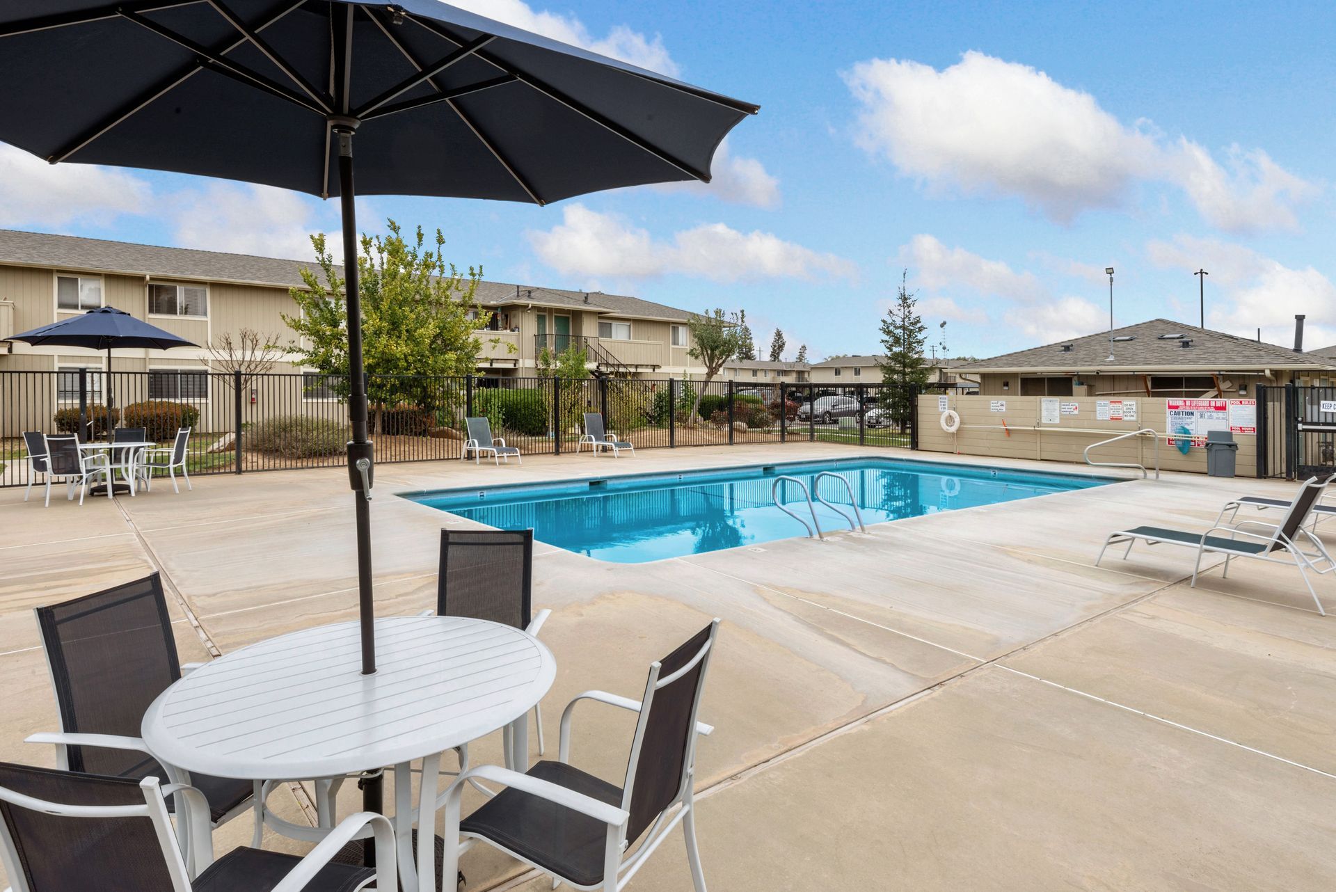 Pool area with umbrella, table, chairs, and apartments in the background. Blue water, sunny day.
