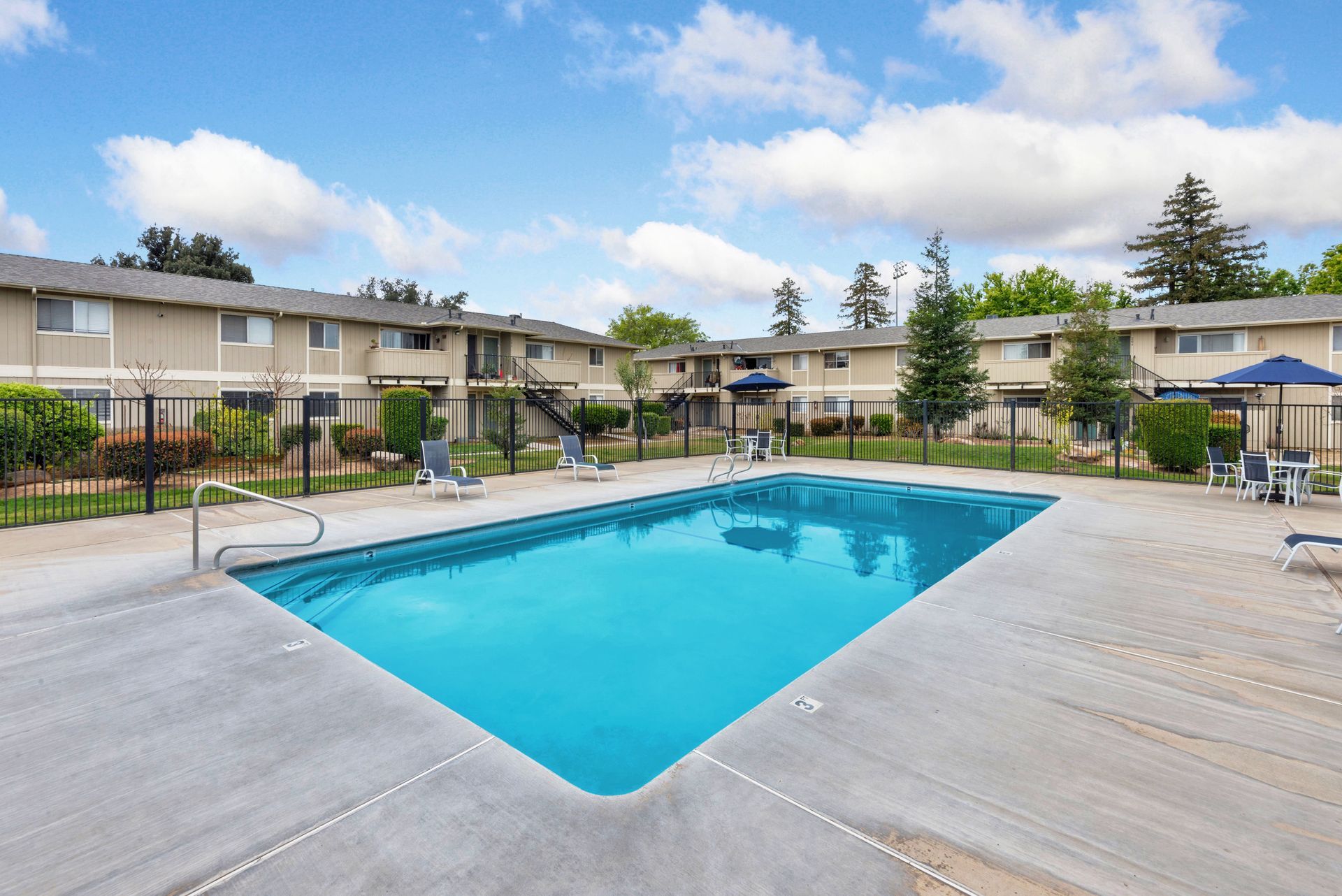 Swimming pool in a courtyard surrounded by two-story apartments and greenery. Blue water, concrete deck.