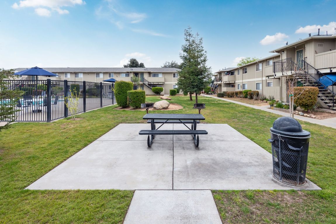 Picnic table and trash can on a concrete slab in a grassy area, apartment buildings in the background.