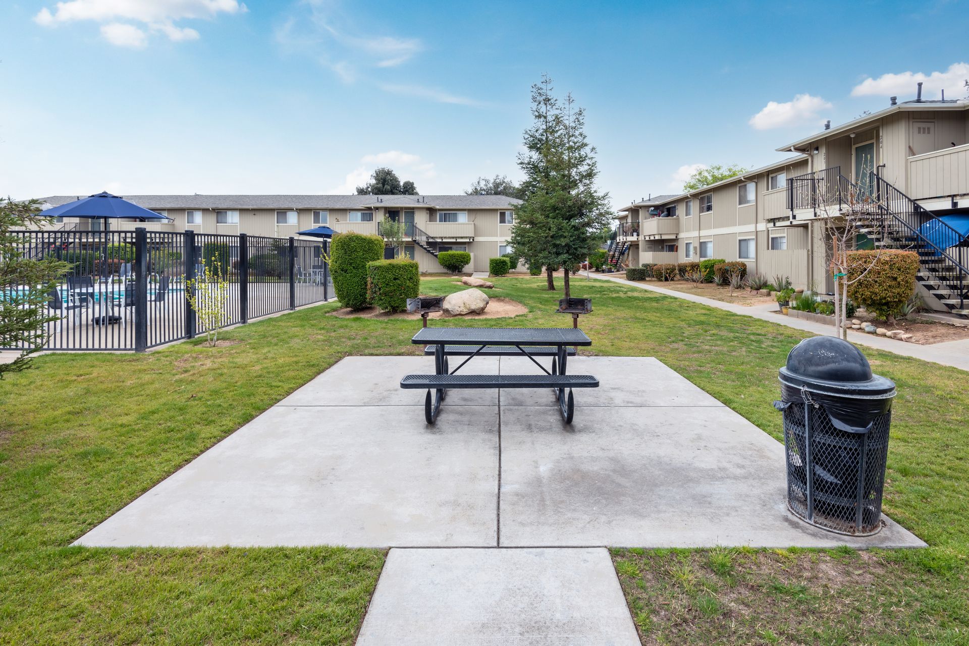 Picnic table and trash can on a concrete slab in a grassy area, apartment buildings in the background.