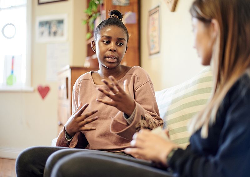 Woman gesturing while talking to another woman on a couch in a living room.