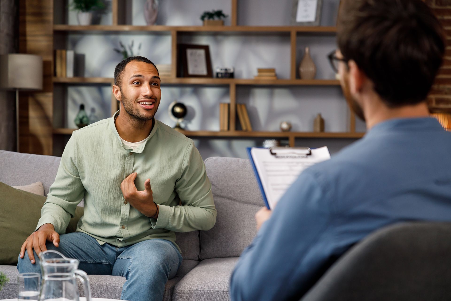 Man in therapy session talking to therapist, sitting on a couch. The therapist is taking notes.