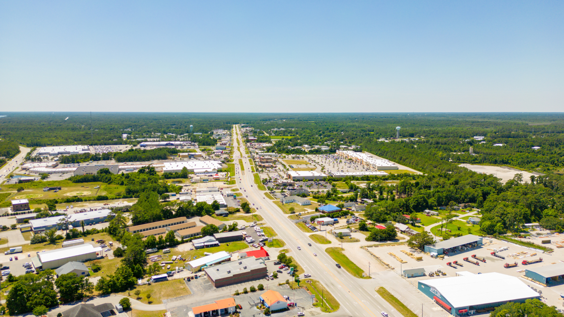Aerial view of a sunny town with a main road lined with buildings, trees, and blue sky.