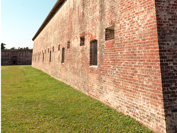 Long brick wall with small windows, curving along a grassy field. Sunlight casts shadows.