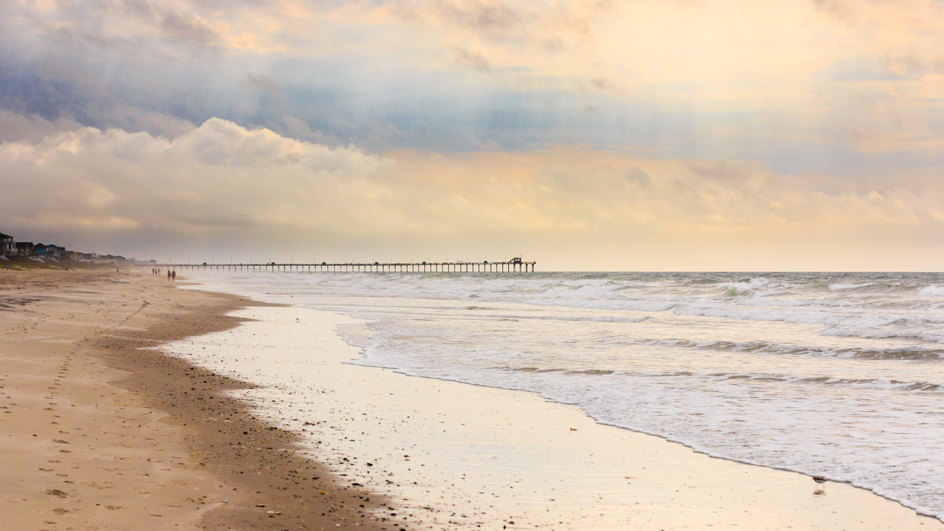 Beach with a long pier extending into the ocean under a cloudy, golden sky. Waves gently roll onto the sandy shore.