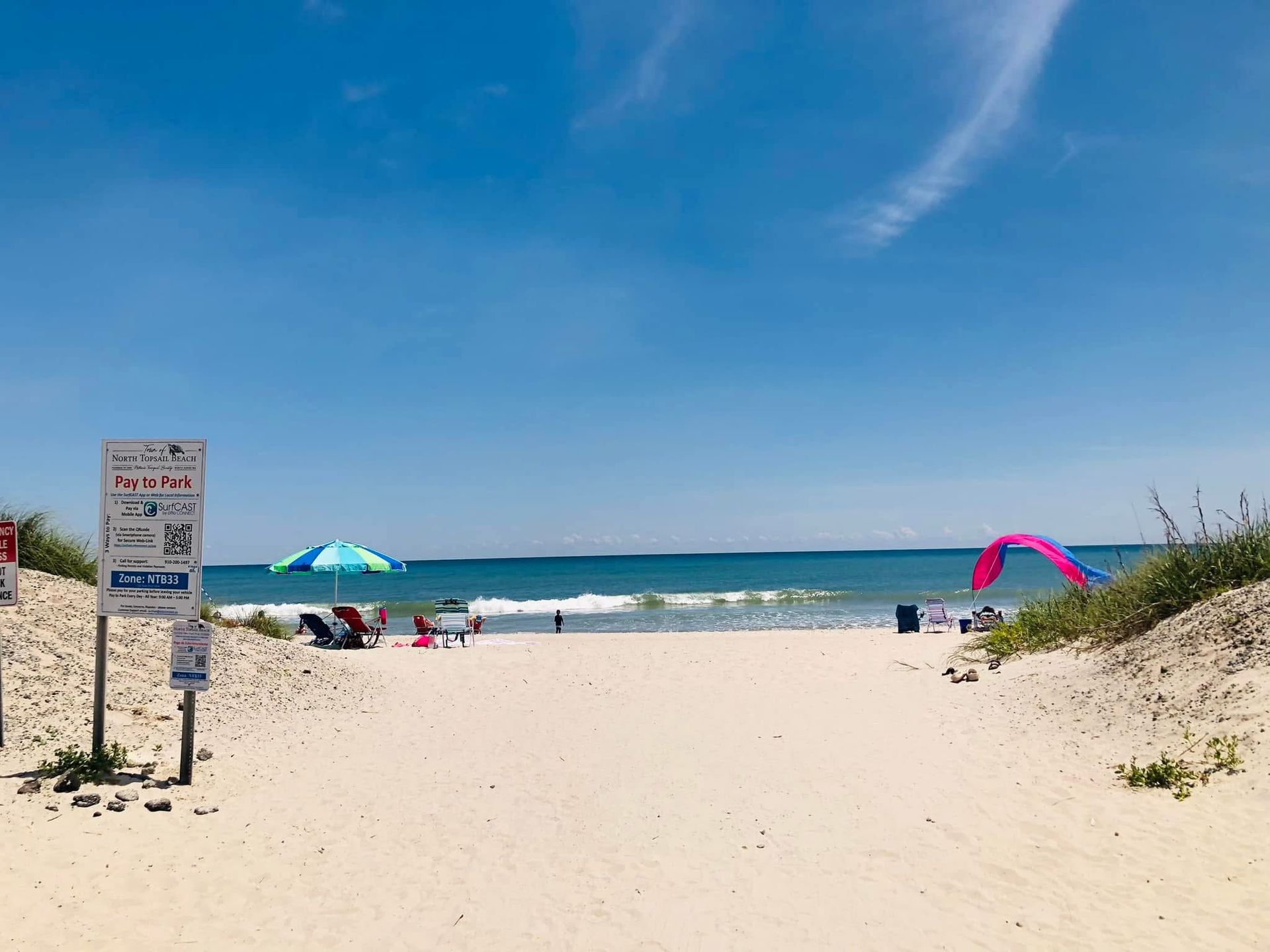 Aerial view of a beach with turquoise water, white sand, and many blue umbrellas. Houses line the shore.