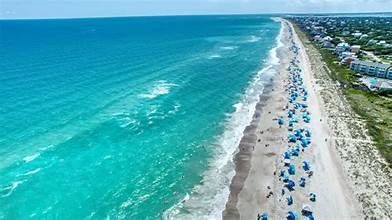 Aerial view of a beach with turquoise water, white sand, and many blue umbrellas. Houses line the shore.