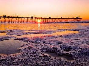 Sunset over ocean with pier; orange sky, foamy water.