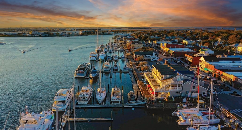 Harbor at sunset with docked sailboats and buildings. Orange and blue sky reflects on water.
