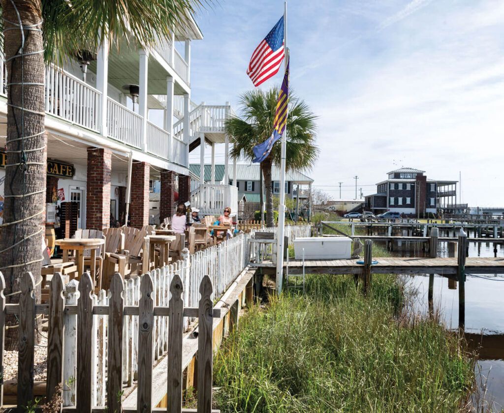 Restaurant with outdoor seating, waterfront view.  American flag waves. White fence, water, building with balconies.