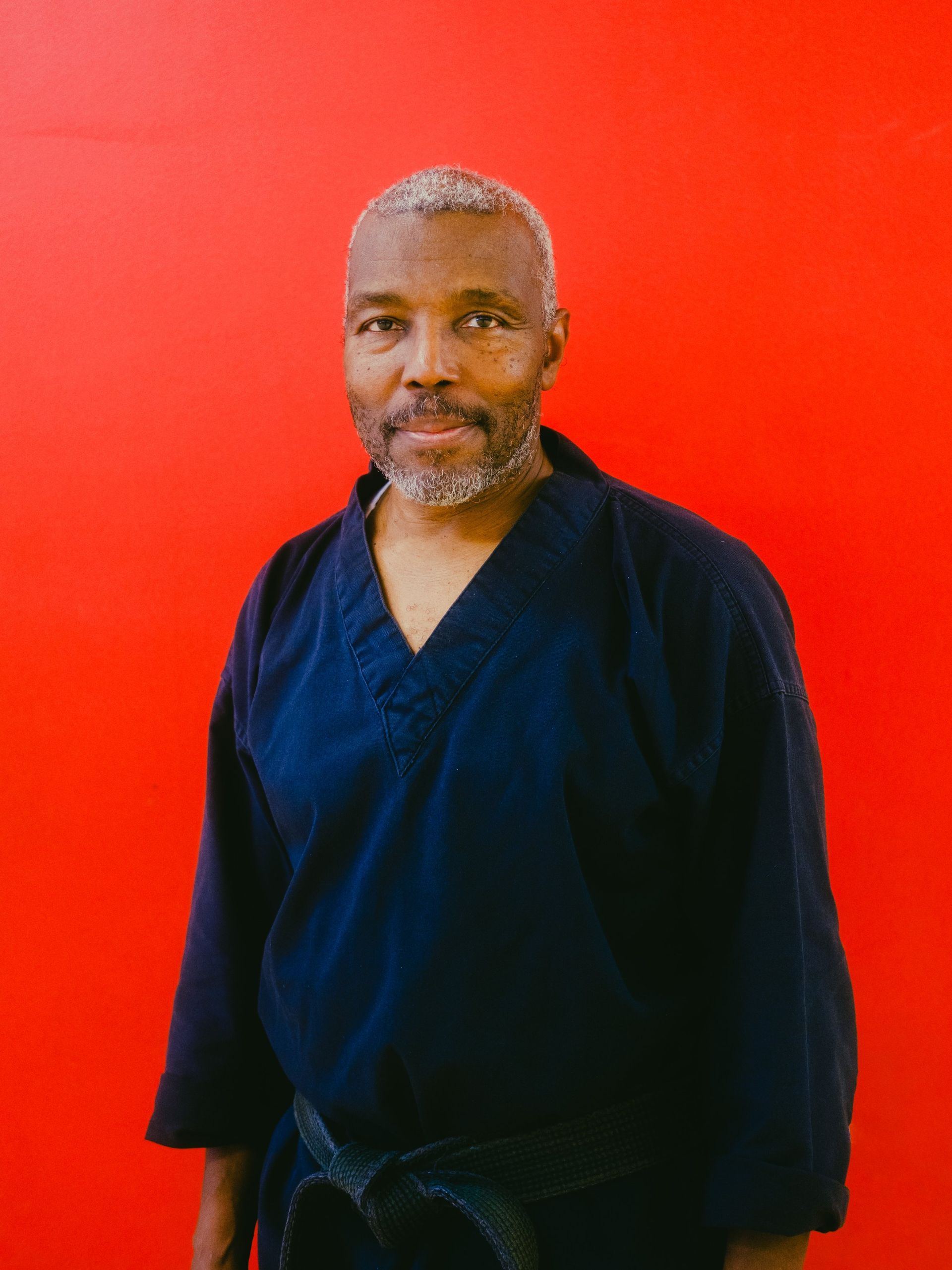 A man in a black karate uniform is standing in front of a red wall.