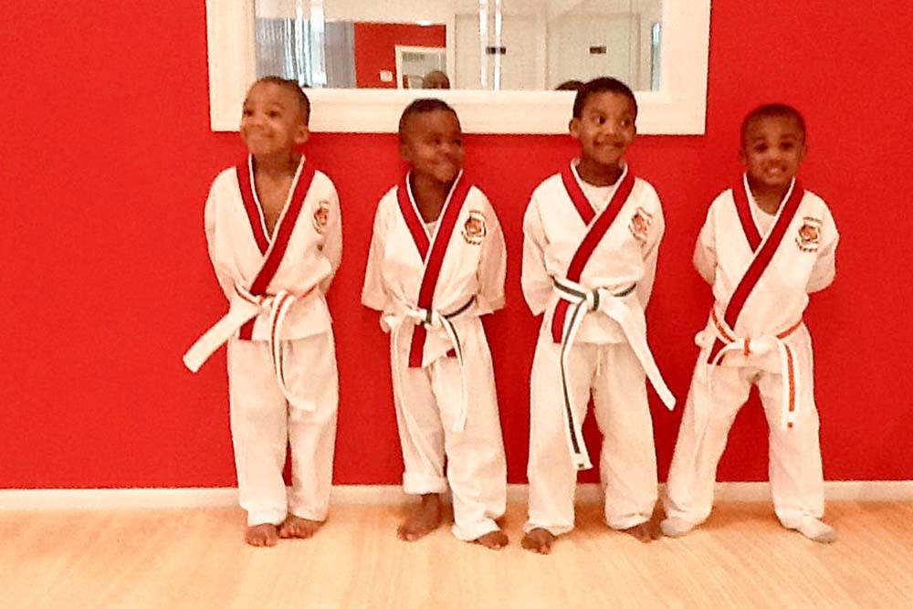 Four young boys in karate uniforms are standing in front of a red wall.