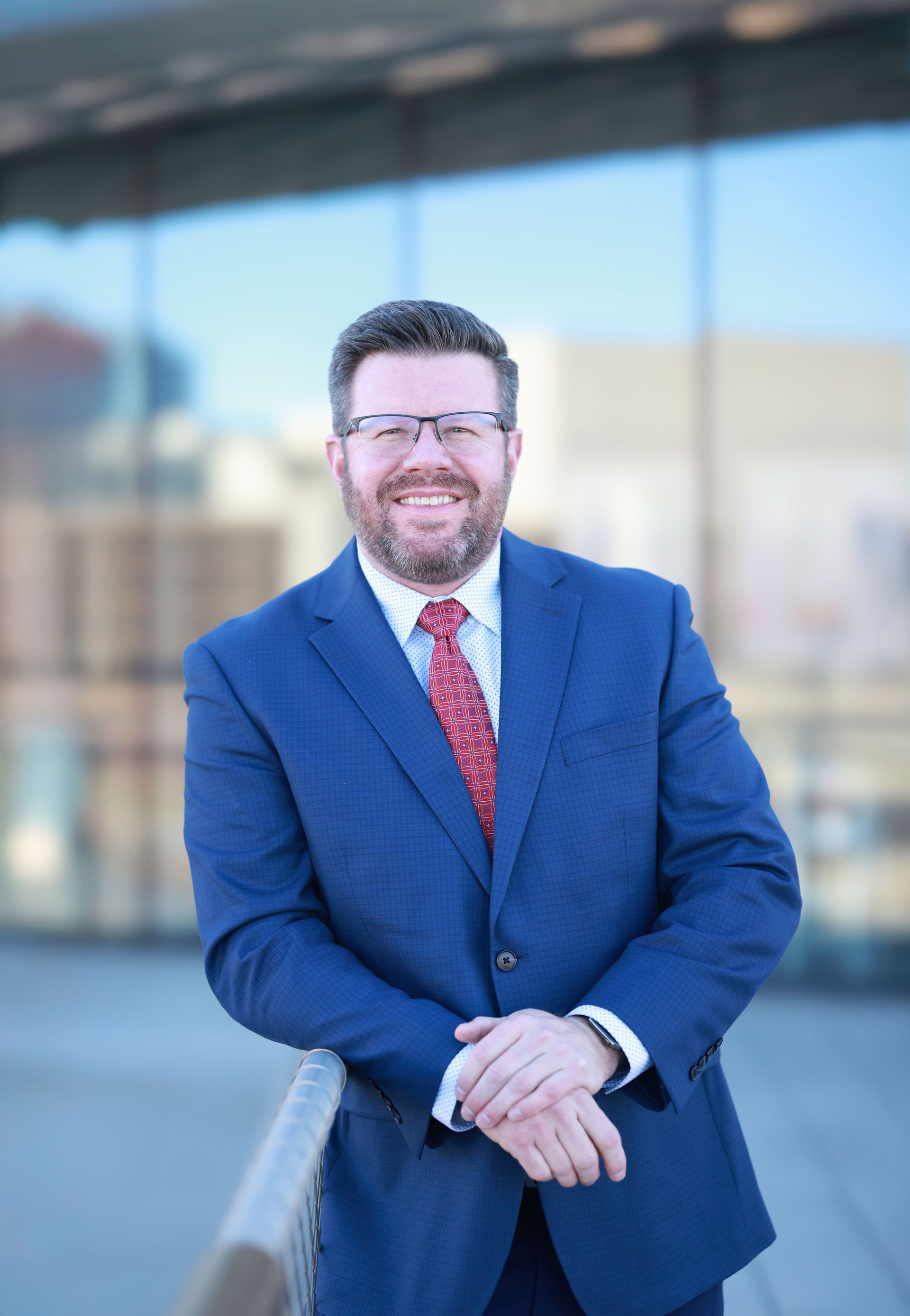Michael Peck  in a blue suit and red tie is standing in front of a building.