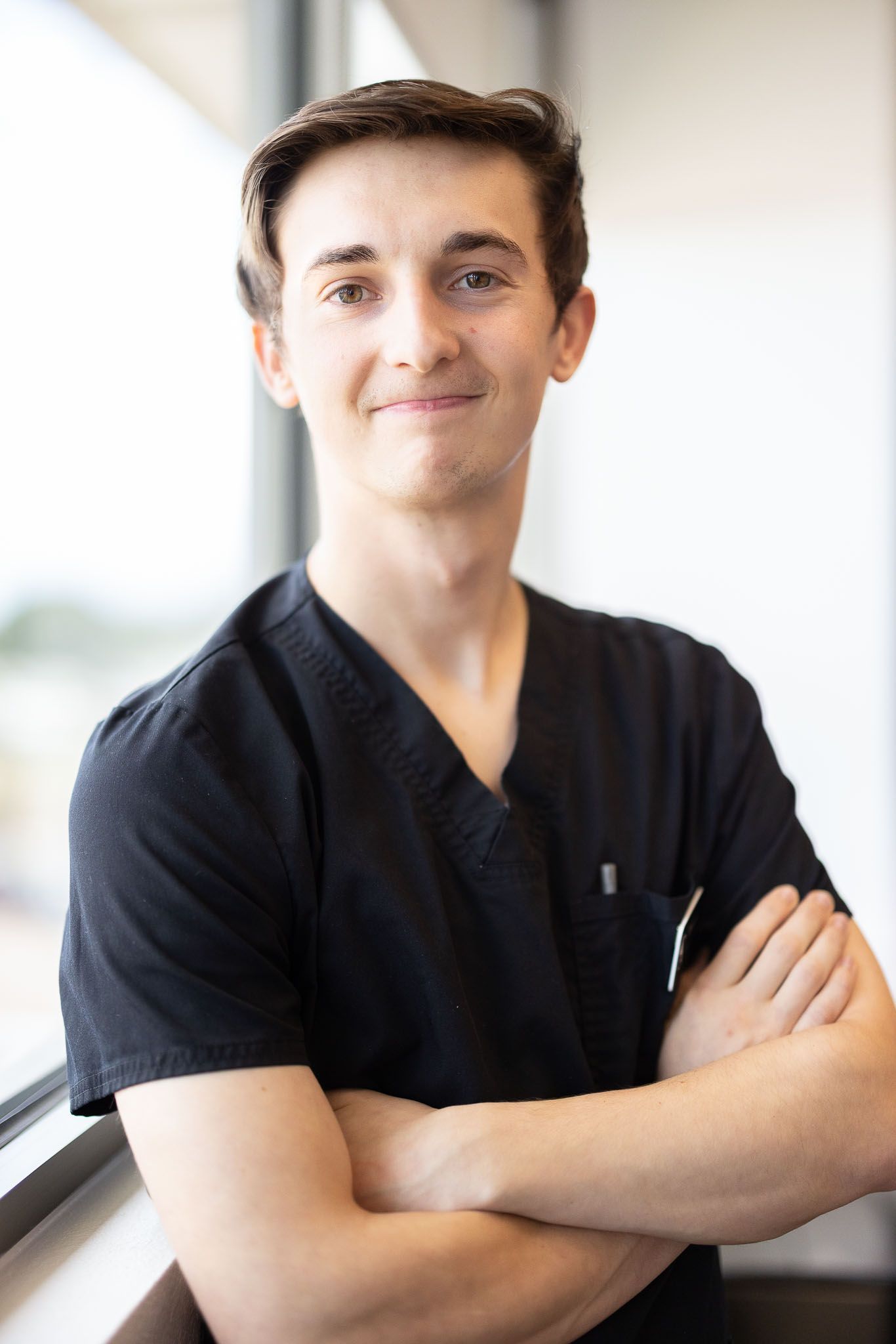 Dennis the Medical Assistant is in a black scrub top is smiling with his arms crossed.