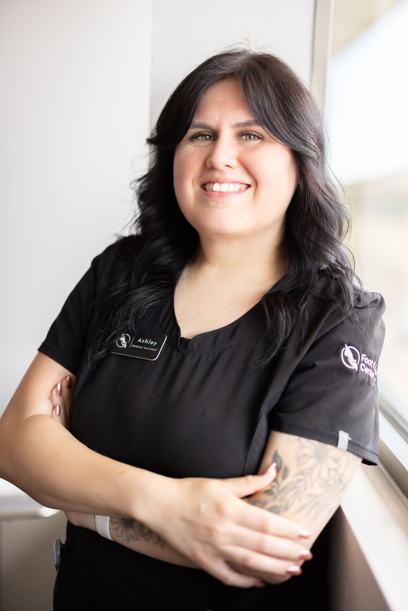 Ashley the Medical Assistant is in a black scrub top is smiling with her arms crossed.