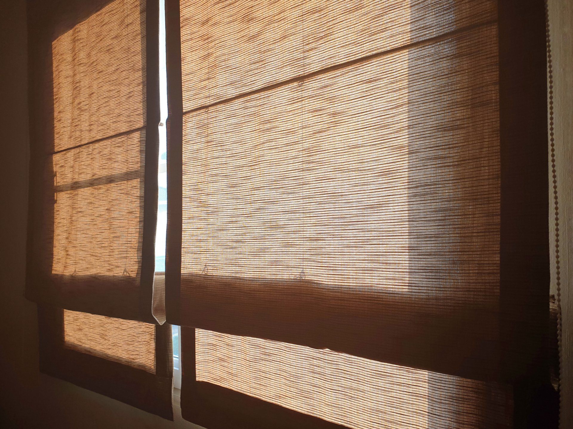Brown woven Roman shade partially covering a window, casting shadows.