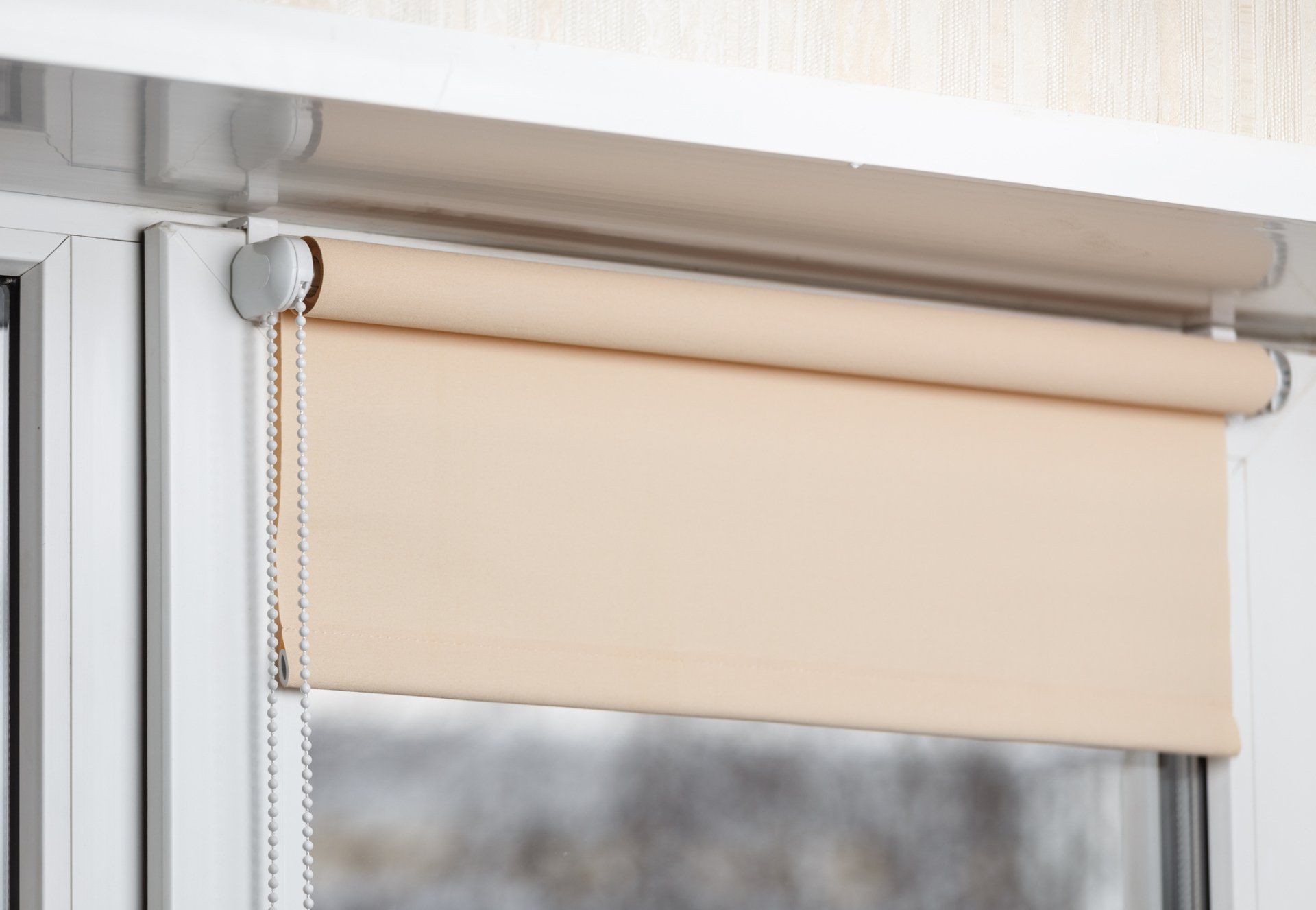 Beige roller blind partially covering a window, with a beaded chain on the left.