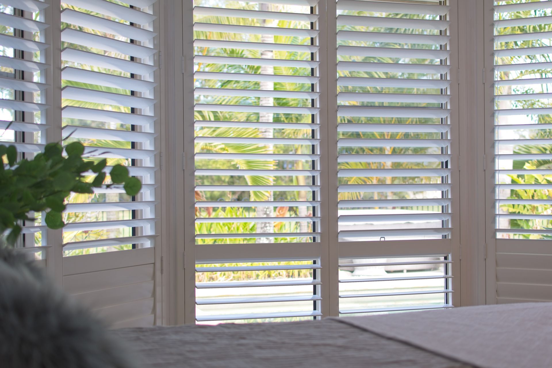Luxury white indoor plantation shutters in a bedroom with green foliage outdoors.
