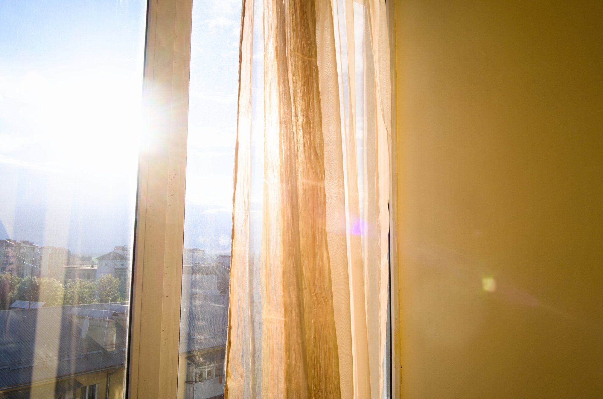 Sunlight streams through a window with sheer gold curtains, illuminating an urban cityscape.