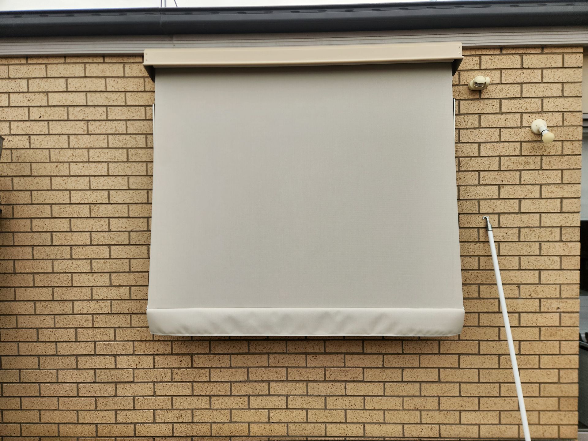 Beige outdoor roller blind on a brick wall, partially lowered, with a white pole.