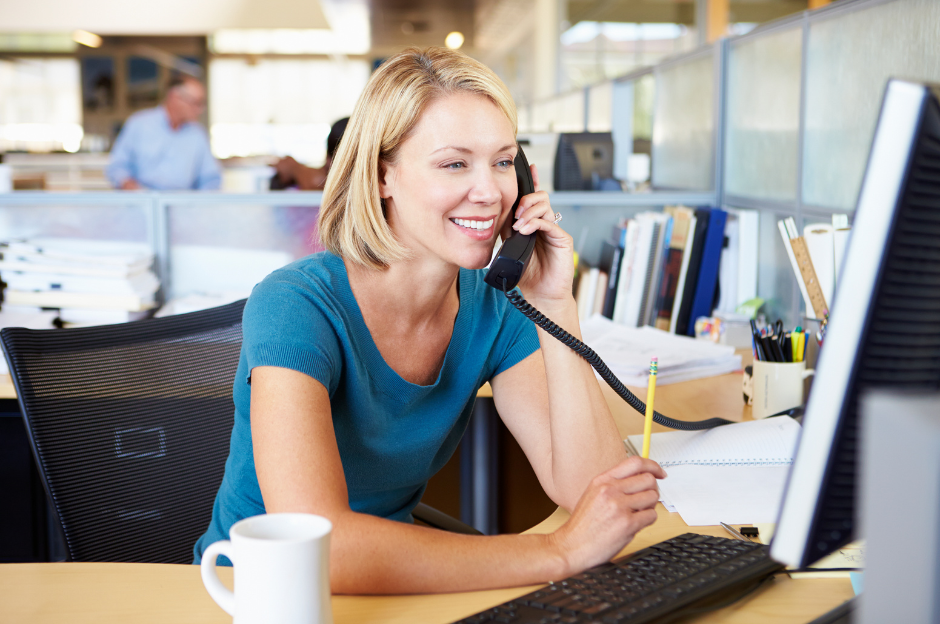 Woman smiling while talking on the phone in an office, looking at computer, pencil in hand, cup nearby.